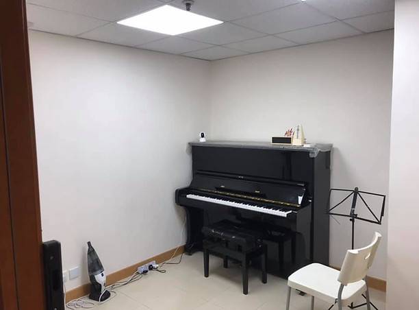 一間小而整潔的音樂練習室，白色的牆上放著一架黑色的直立式鋼琴，一個樂譜架，一張椅子，角落裡有一個吸塵器。