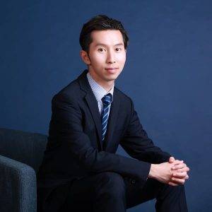 一位身著深色西裝、打著條紋領帶的男子坐在藍色背景的椅子上，雙手緊握，面對鏡頭。.