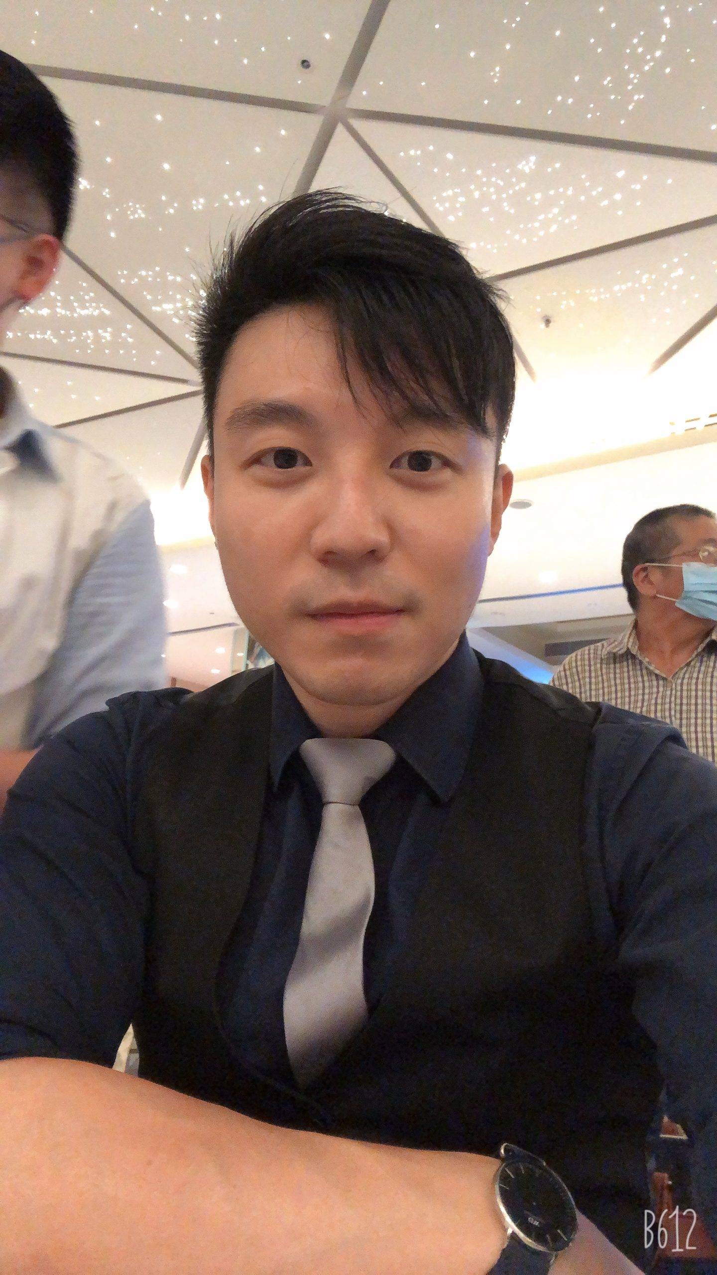 一名穿著深色襯衫、背心和領帶的男子在室內看著攝影機，背景是另外兩個人，其中一人戴著口罩。.