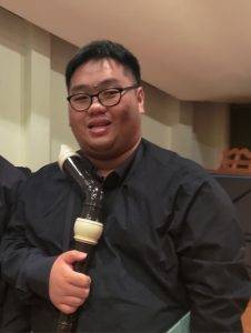 一位戴著眼鏡、身穿黑色襯衫的人拿著一台大型黑色錄音機，站在室內靠著一堵米色牆壁。.