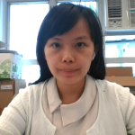 一位黑色直髮的女士坐在室內的辦公桌前，望著鏡頭。背景可見紙箱和冷氣機。.