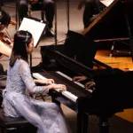 一位身穿藍色禮服的年輕女性在舞台上與樂團一起彈奏三角鋼琴；UKMEA 的標誌顯示在右上角。.