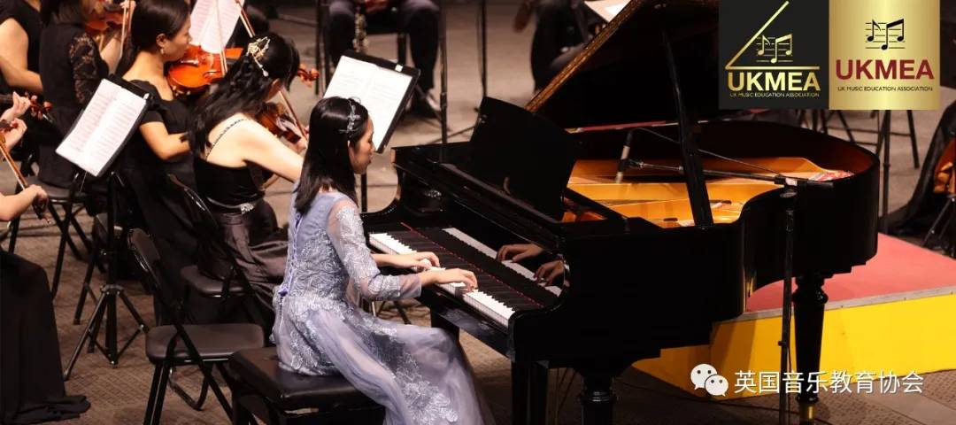 一位身穿藍色禮服的年輕女性在舞台上與樂團一起彈奏三角鋼琴；UKMEA 的標誌顯示在右上角。.