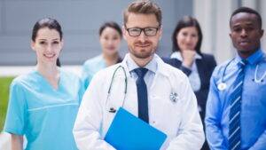 一群包括醫生和護士在內的五位醫療照護專業人員，穿著醫療服裝站在戶外，看著攝影機。.