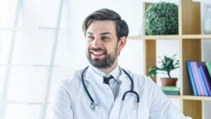 一位穿著白大褂、戴著聽診器的男醫生坐在明亮的辦公室裡微笑，背景是架子和植物。.