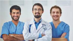 三位身穿藍色手術服和白大褂的男性醫療專業人員站在一起，在臨床環境中對著鏡頭微笑。.