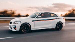 日落時分，一輛帶有賽車條紋的白色 BMW 轎跑車在高速公路上行駛，背景為動態模糊影像。.