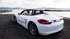 一個人坐在停在水體附近的白色敞篷跑車裡，背景是群山和多雲的天空。.