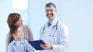 在明亮的醫療辦公室裡，一位拿著記事板的醫生微笑地站在一位女士和一位年輕女孩身旁，兩人都在看著他。.