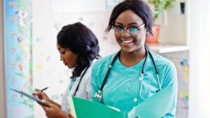 兩位穿著手術服的專業醫護人員站在醫療環境中，其中一位拿著文件夾對著鏡頭微笑，另一位則在背景的剪貼板上寫字。.