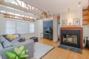 現代化客廳，天花板橫樑外露、灰色組合沙發、壁爐、電視、木地板、地毯，角落還有一盆大植物。.