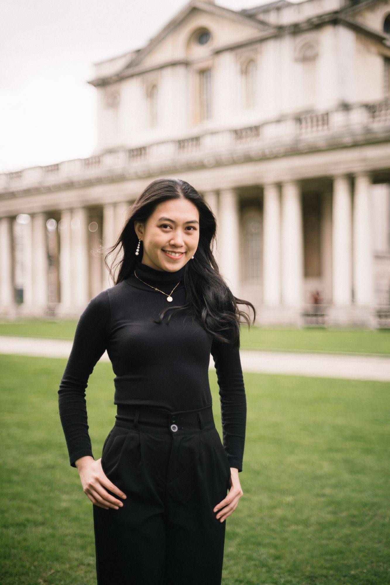一位身穿黑色上衣和褲子的女士微笑地站在一座有圓柱的歷史建築前的草坪上。.