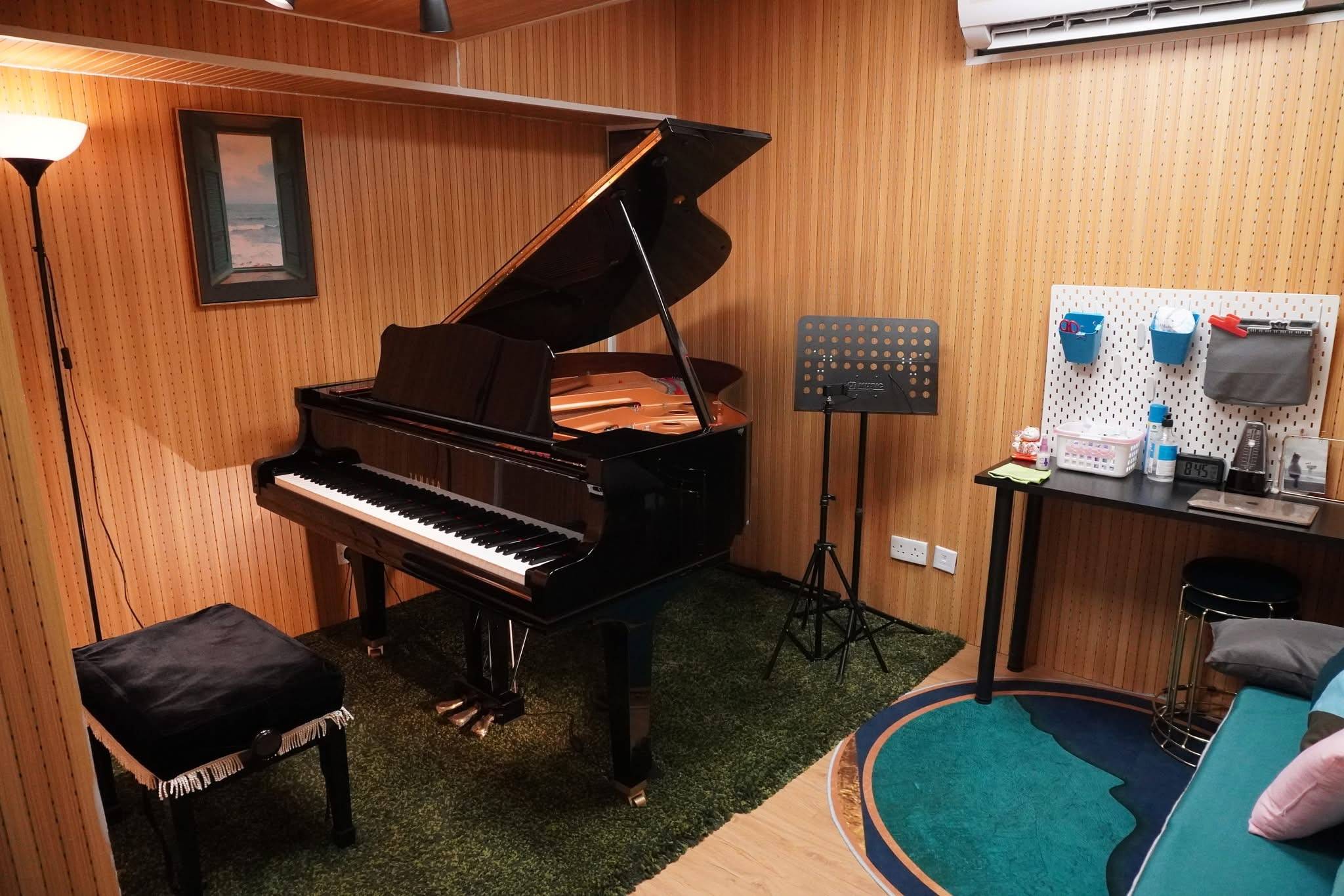 在一個舒適的房間裡，放置著一架附有長椅的三角鋼琴，房間牆壁以木質鑲板鋪成，並鋪有地毯、譜架、書桌和空調。.
