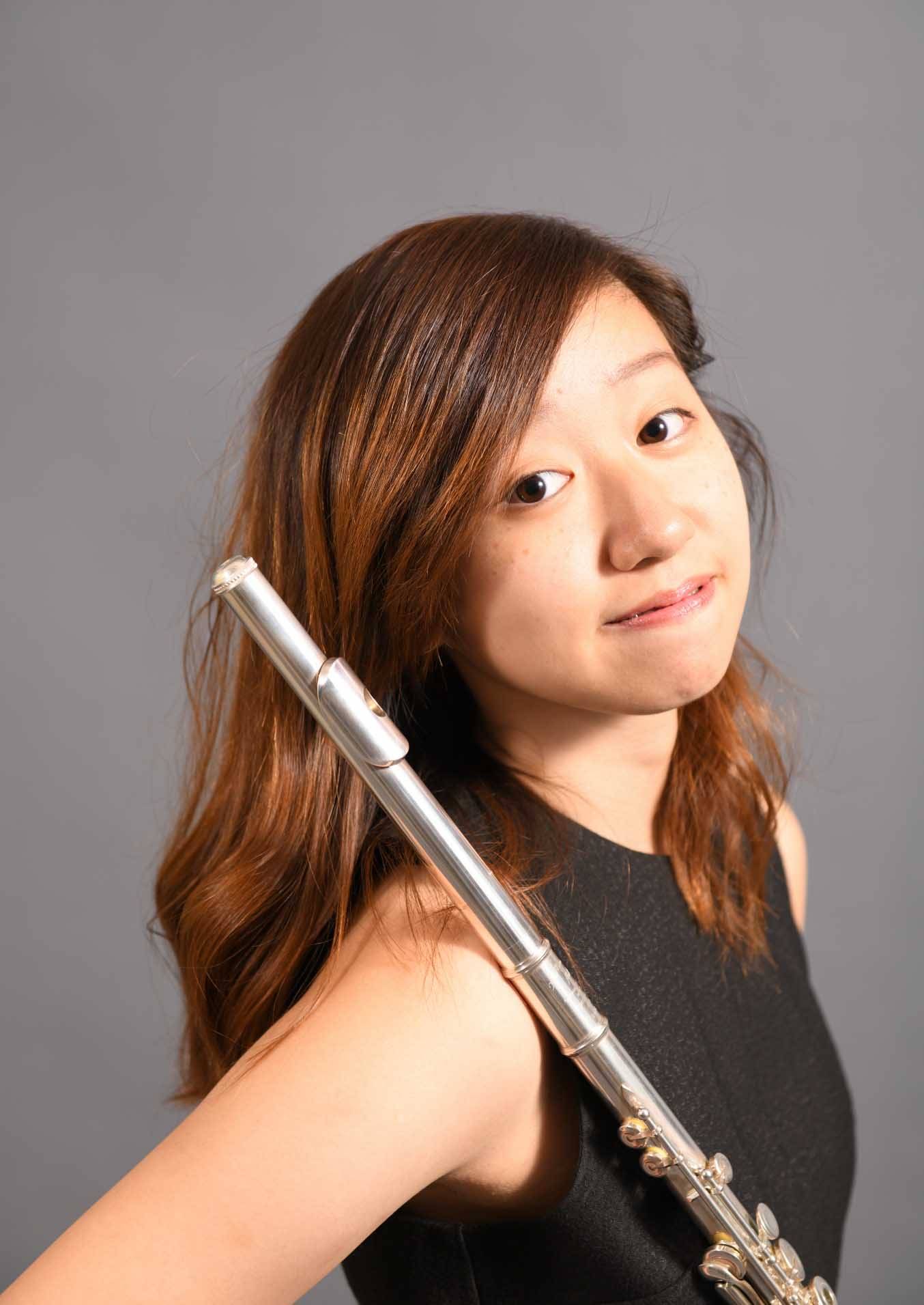 一位棕色長髮的女性肩上橫握著一支銀色長笛，在素雅的灰色背景中望著鏡頭。.