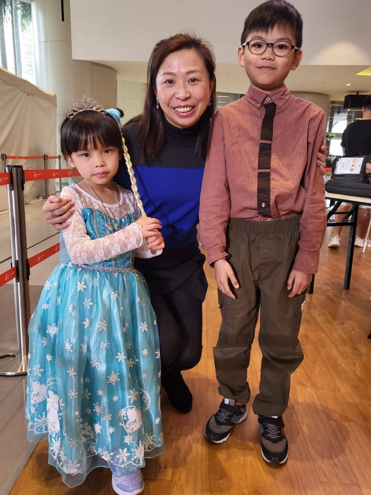 一位女士在室內與兩個孩子合照；女孩穿著藍色公主裙和頭飾，男孩則戴著眼鏡，穿著棕色襯衫搭配綠色褲子。.