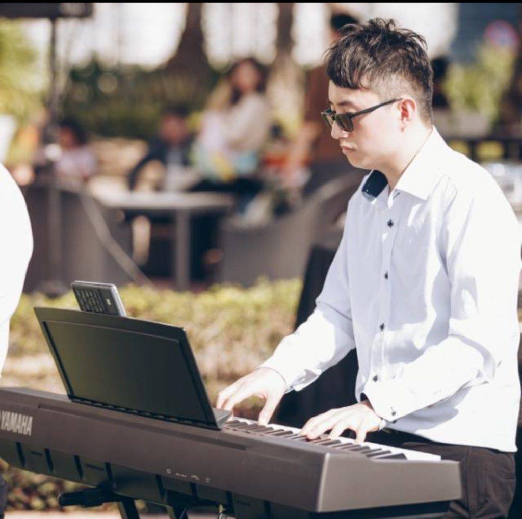 一個穿著白襯衫的人在戶外演奏 Yamaha 鍵盤，背景是模糊的人和綠色植物。.
